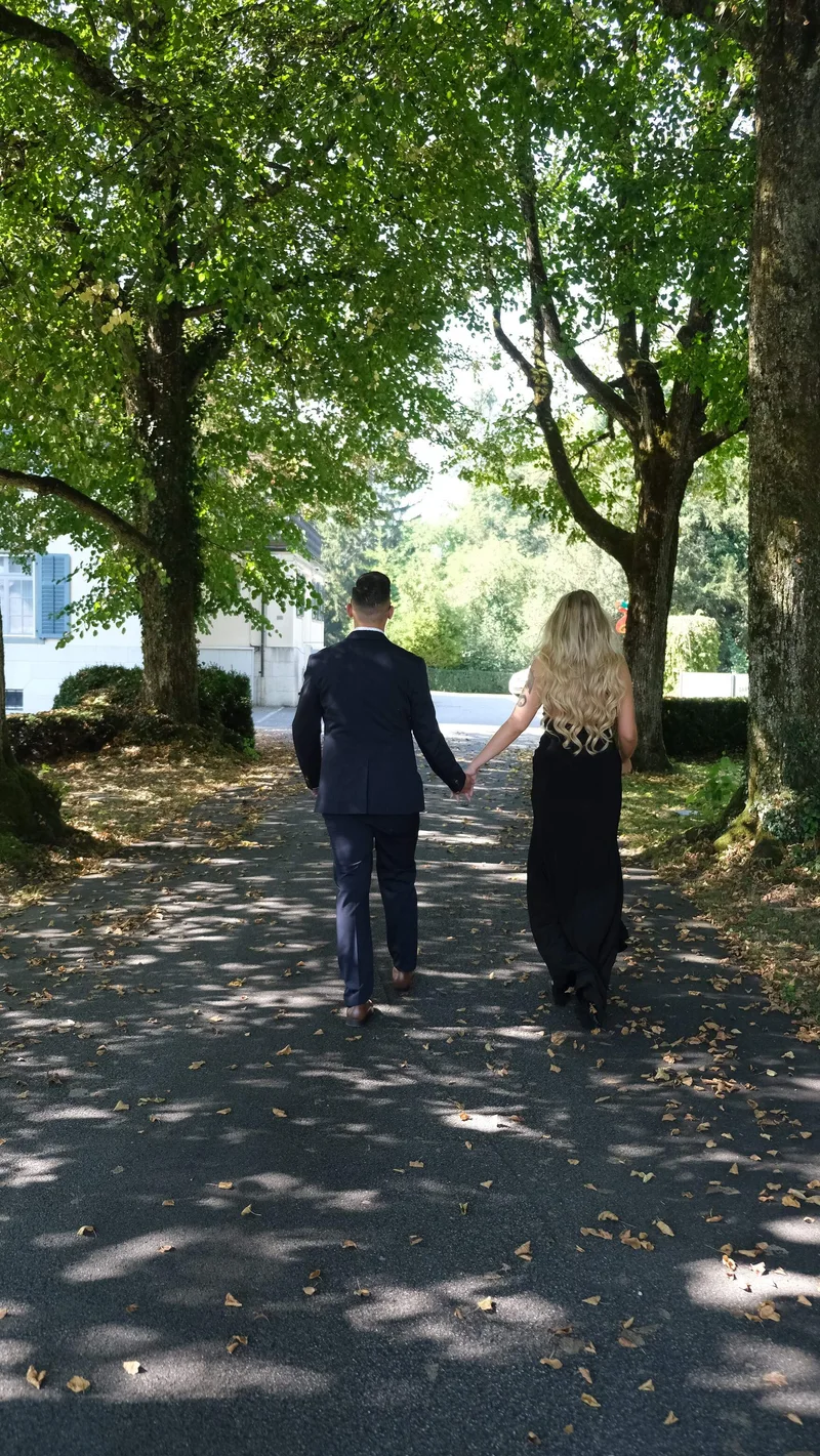 Zwei Menschen gehen Hand in Hand auf einem Weg unter grünen Bäumen.